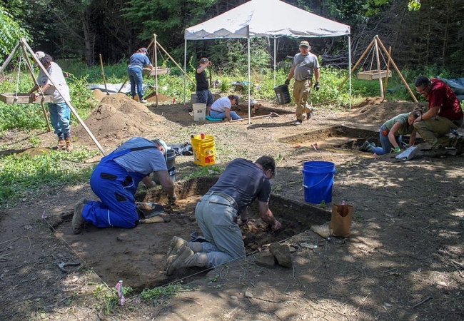 Several people are bent over dig sites or sifting dirt at an archaeological dig.