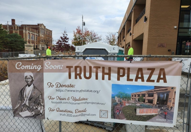 A banner hanging on a wire fence with the title, "Coming Soon: Truth Plaza."