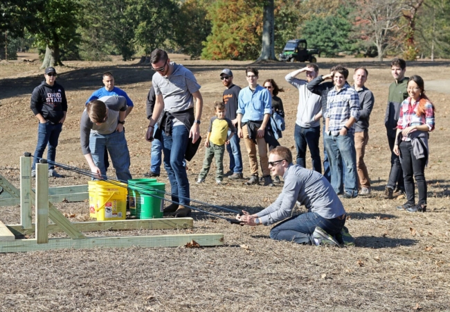 A volunteer pulls back a large slingshot preparing to launch tree nuts into the park while a group of volunteers watches.