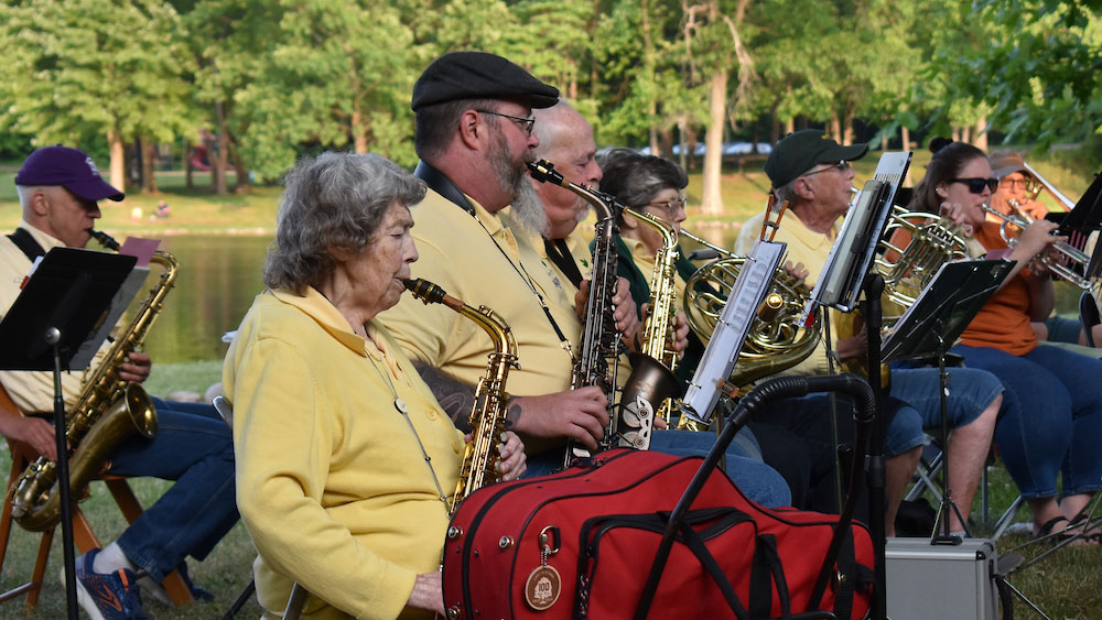 people playing instruments with green trees and a lake in the background on a sunny day