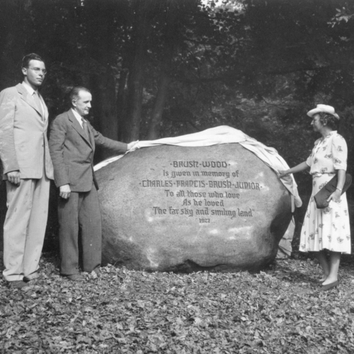 A black and white photo showing a large boulder with etched words. A woman stands to the right of the boulder and two men to the left. The boulder reads, "Brushwood is given in memory of Charles Francis Brush Junior to all those who loved as he loved the far sky and smiling land. 1927"