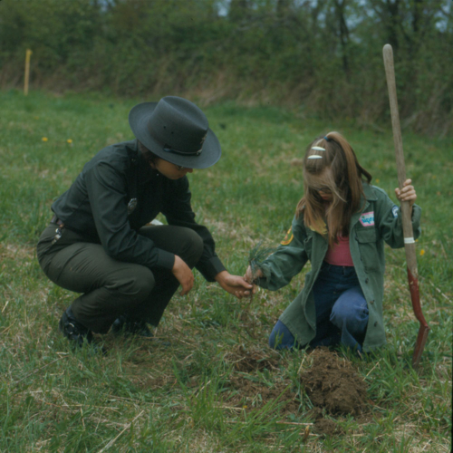 A woman in a ranger uniform is helping a young girl with a shovel plant a tree sapling.