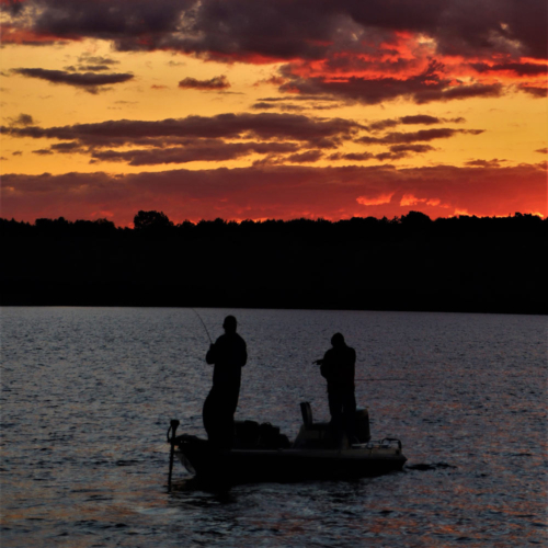 The silhouette of two people fishing from a boat at dusk.