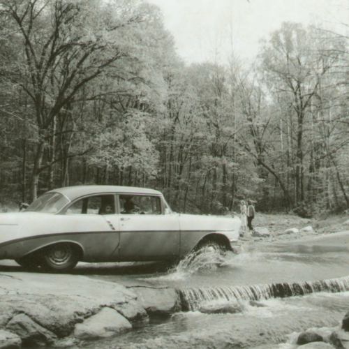 A black and white photo of a car driving through the water of a ford.