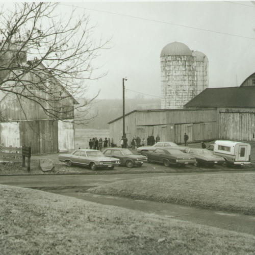 A black and white photo of several cars parked outside of a barn.