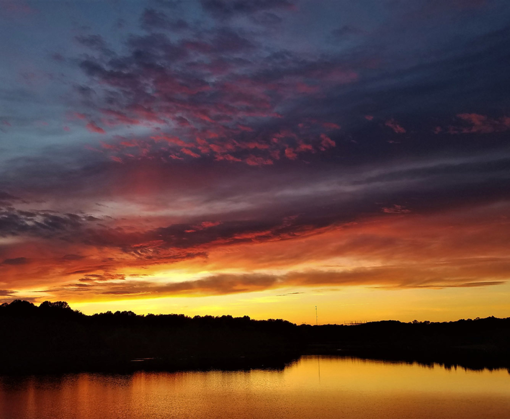 A tree lined lake with a purple, pink and yellow sky above.